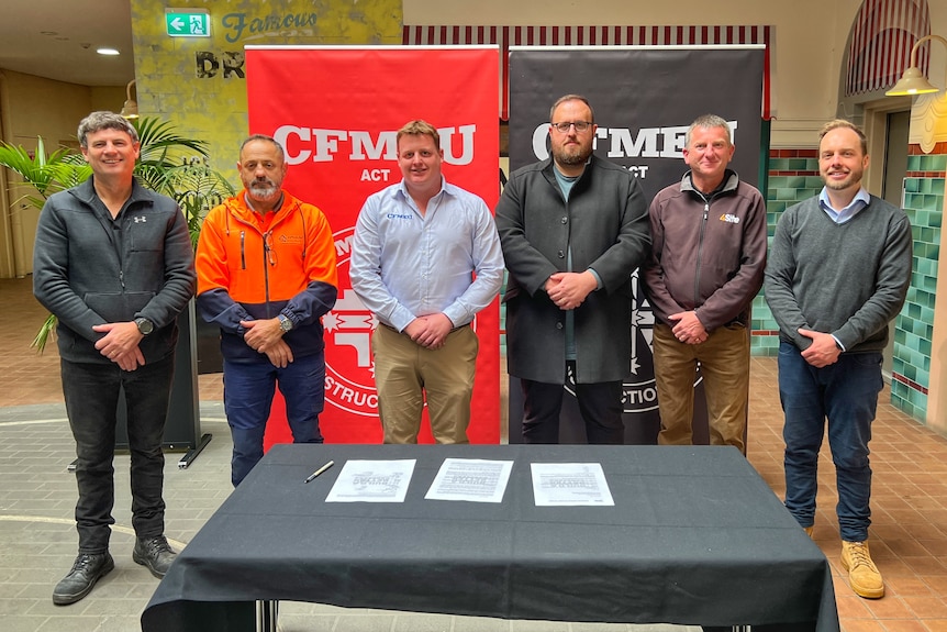 Six men stand in a row in front of signs that say CFMEU.