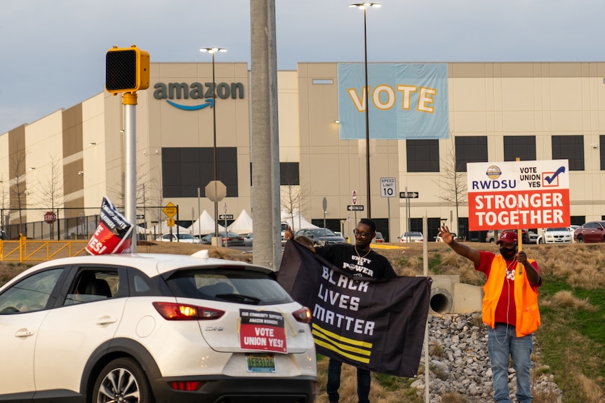 men standing outside of an amazon factory wave pro-union signs at passing cars