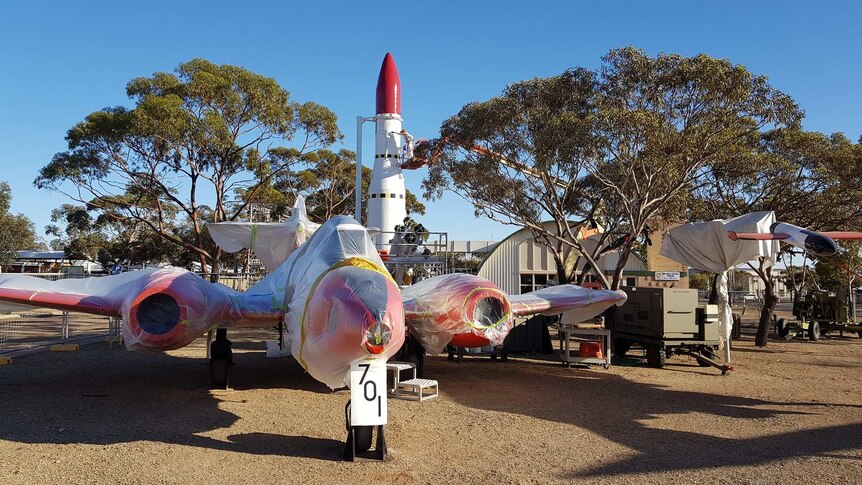 A large white aircraft with red wings is parked on brown dirt in front of a large white missile with a red nose.