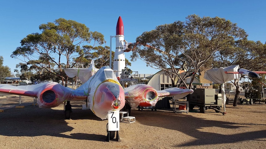 A large white aircraft with red wings is parked on brown dirt in front of a large white missile with a red nose.