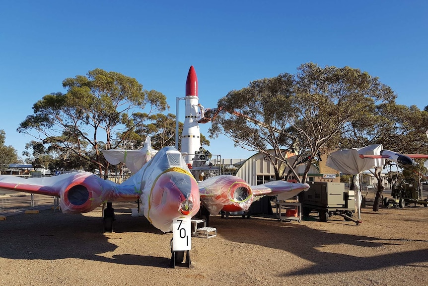 A large white aircraft with red wings is parked on brown dirt in front of a large white missile with a red nose.