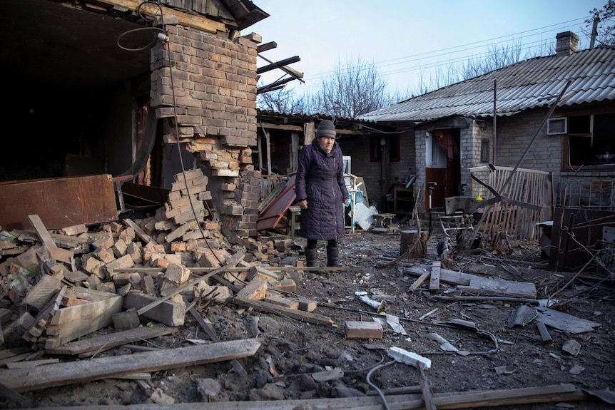 Elderly woman stands next to her damaged house, with piles of bricks on the ground.