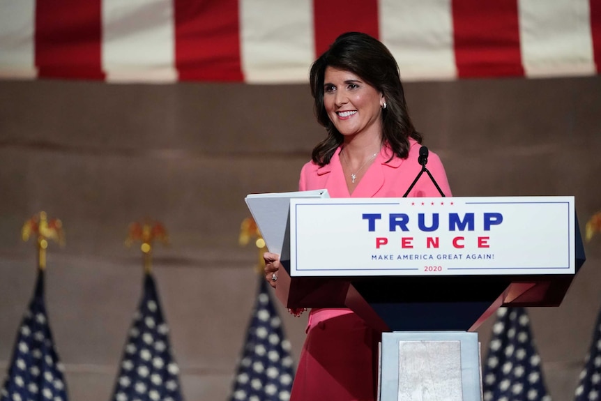 A woman with brown hair and wearing a pink dress stands at a podium with Trump Pence written on it.