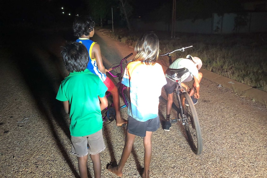 A photo of four children playing on their bikes in the street at night.