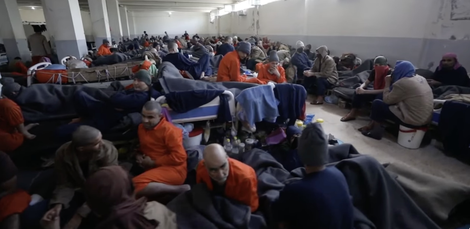 Men in orange prison uniforms sit on matresses in a crowded room