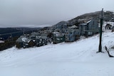 A handful of buildings are seen on a mountaintop. The sky is grey, the ground blanketed in snow and there is a ski lift.
