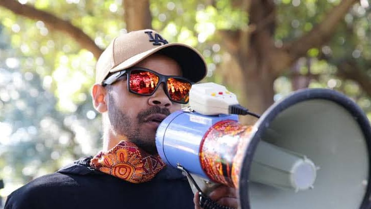 An indigenous young man wearing sunglasses and a hat speaking into a megaphone, branding with indigenous art.