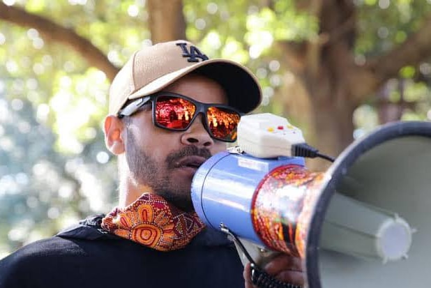 An indigenous young man wearing sunglasses and a hat speaking into a megaphone, branding with indigenous art.