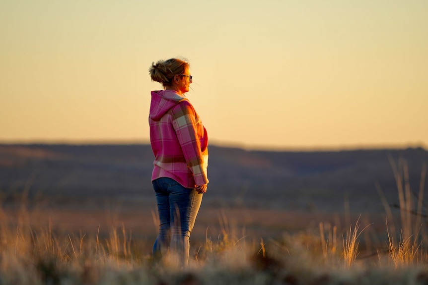A woman stands on a hill and looks towards the sunset.