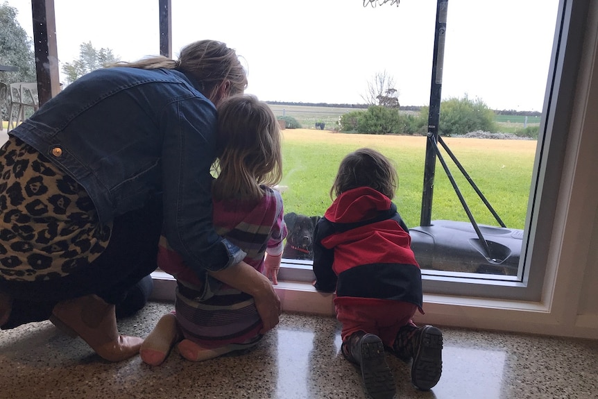 A young mum and two small children look out a window at their house