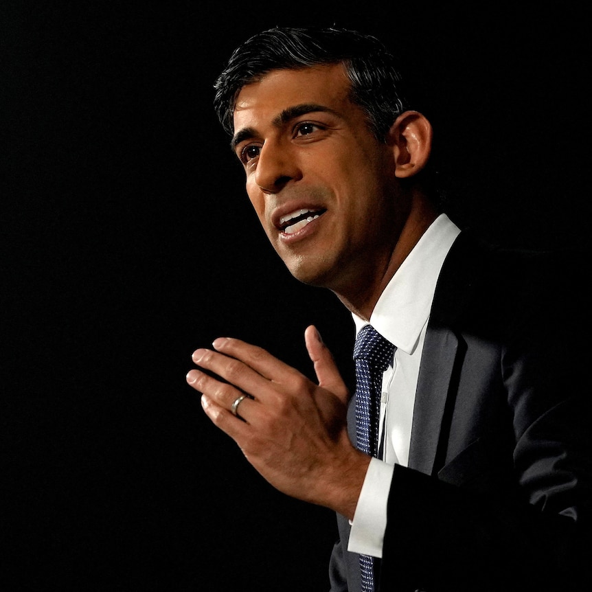 A middle-aged man of Indian heritage in a black suit speaks against a dark backdrop.