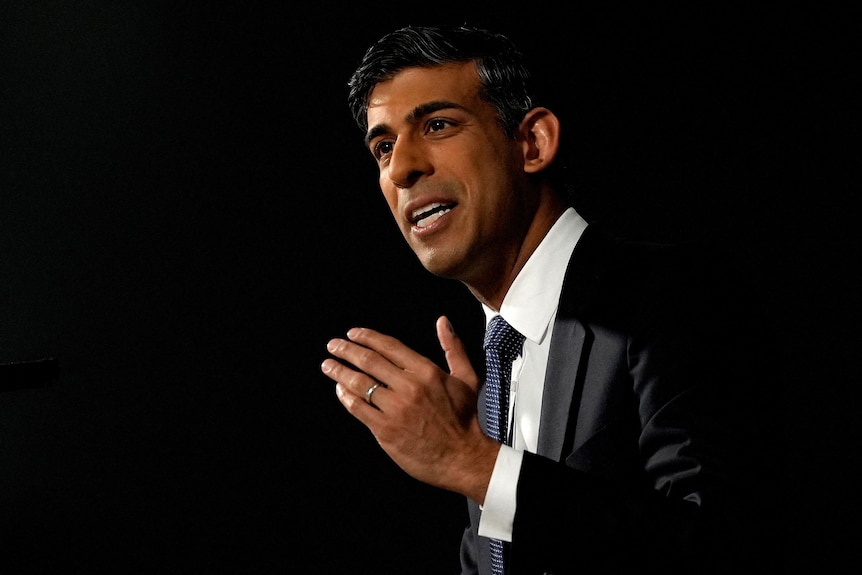 A middle-aged man of Indian heritage in a black suit speaks against a dark backdrop.