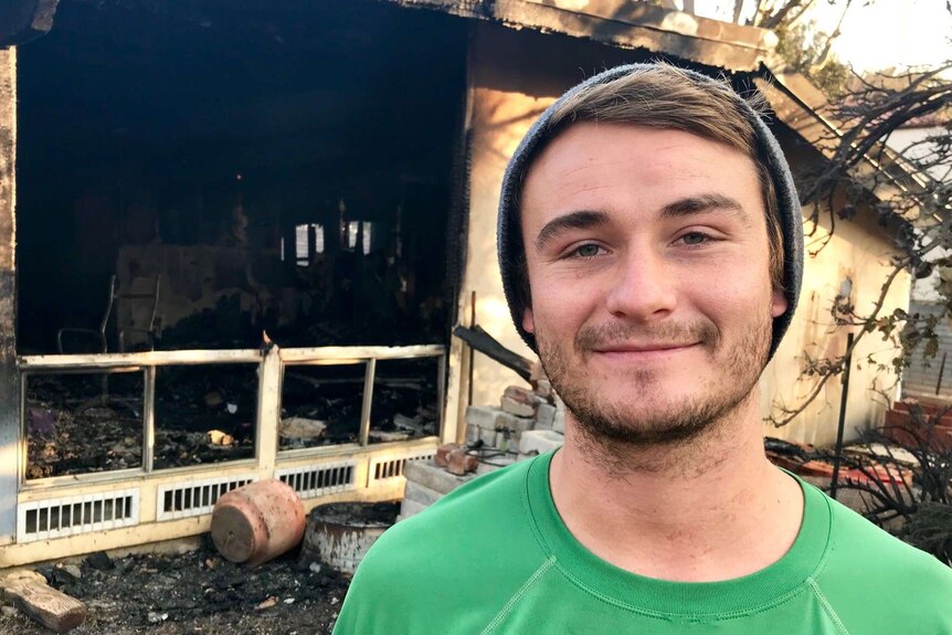 A man wearing a beanie stands in front of a burnt home