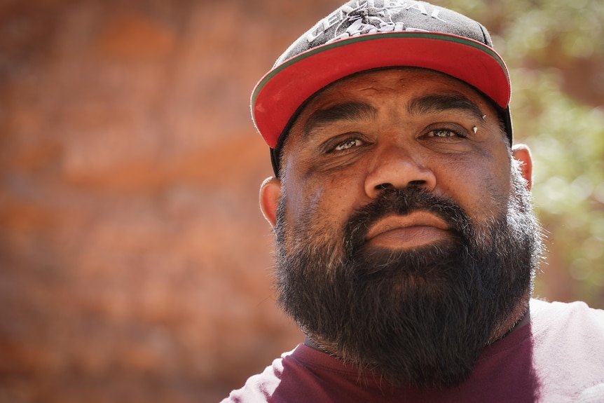 Indigenous man's face close-up, serious, in front of a red rock, black well-groomed beard, red and black cap.