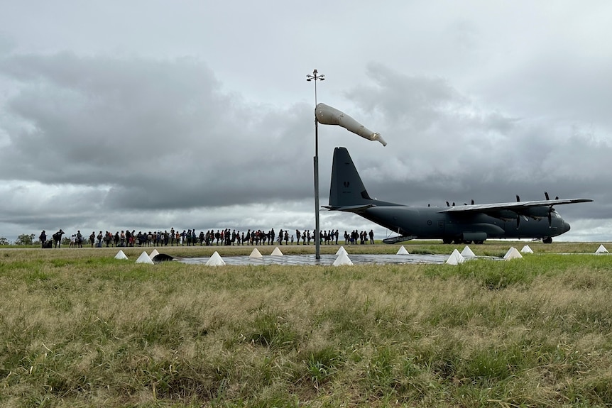 A plane sits on a tar mac with a wind sock in the foreground with a large group of people distant in the background