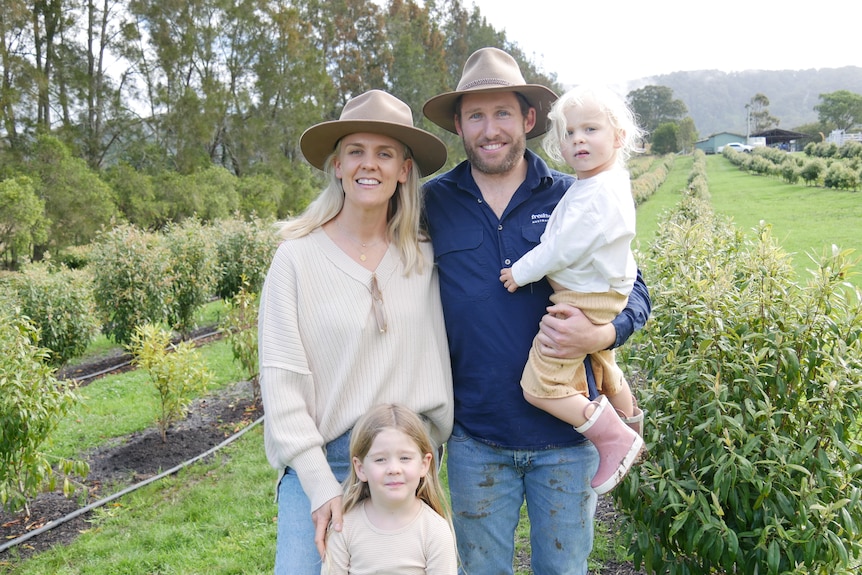 Smiling woman wears hat, light top, jeans, holds child, man in t-shirt, jeans, carries child in front of lemon myrtle plants.