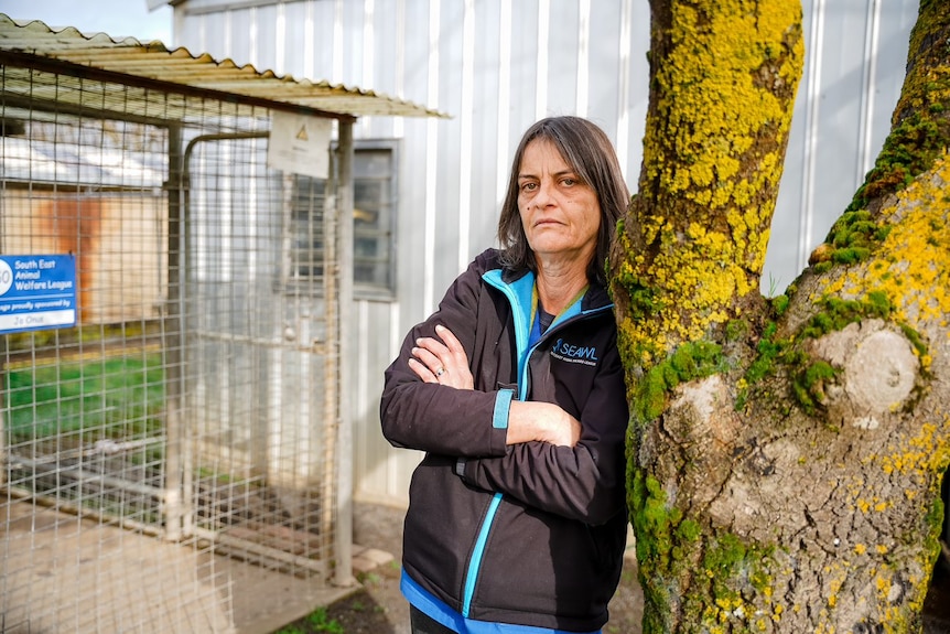 Woman leaning on a tree with arms folded and serious look on face. Dog kennel (animal welfare) in the background.