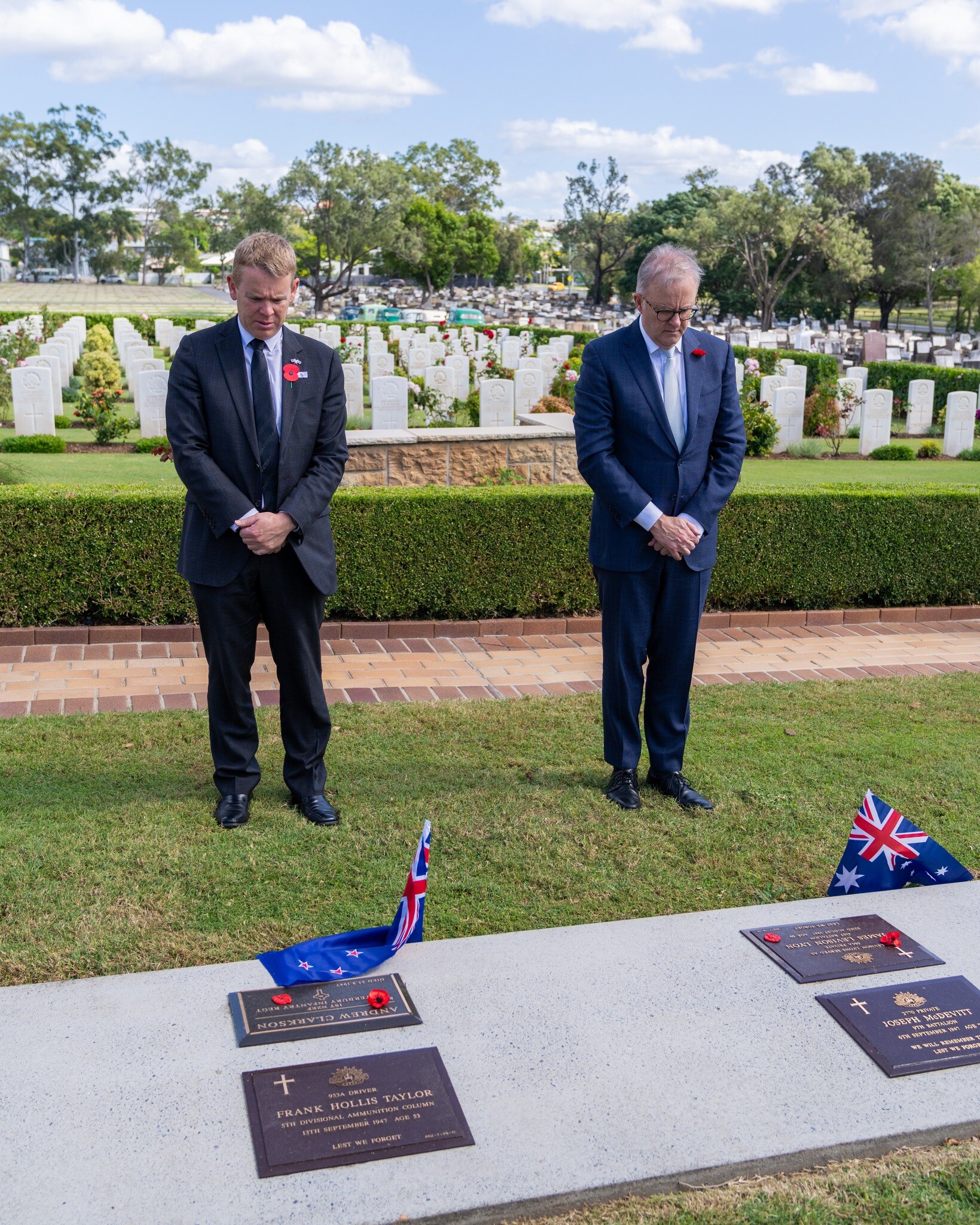 Two men wearing a black and blue suit stand bowing heads in a moment of silence with grave stones in background