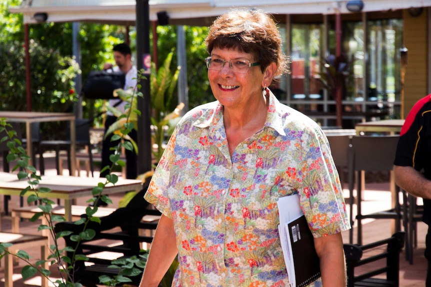 Pat Turner walks towards the camera holding papers and smiling outdoors.