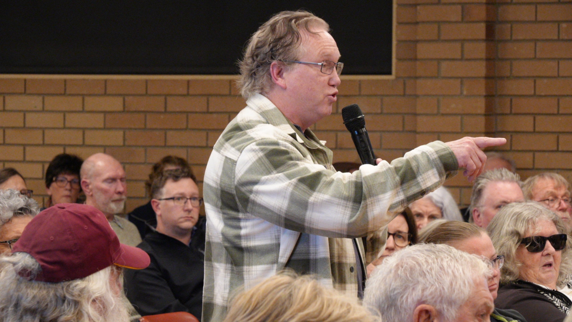A midddle-aged man points as he stands and speaks into a microphone among a crowd in a community hall.