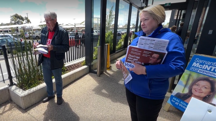 A woman with a blonde bob wearing blue holds how to vote cards on a ramp to a building. There is a carpark in the background