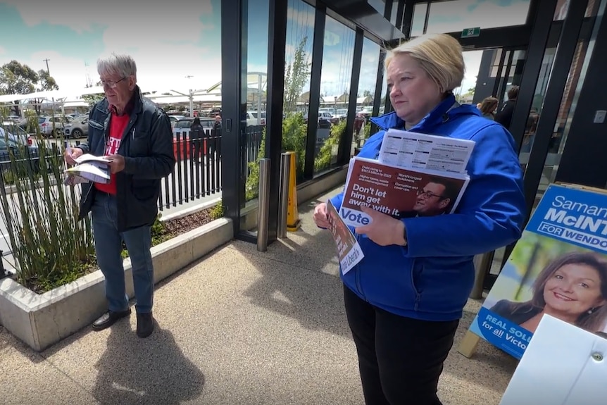 A woman with a blonde bob wearing blue holds how to vote cards on a ramp to a building. There is a carpark in the background