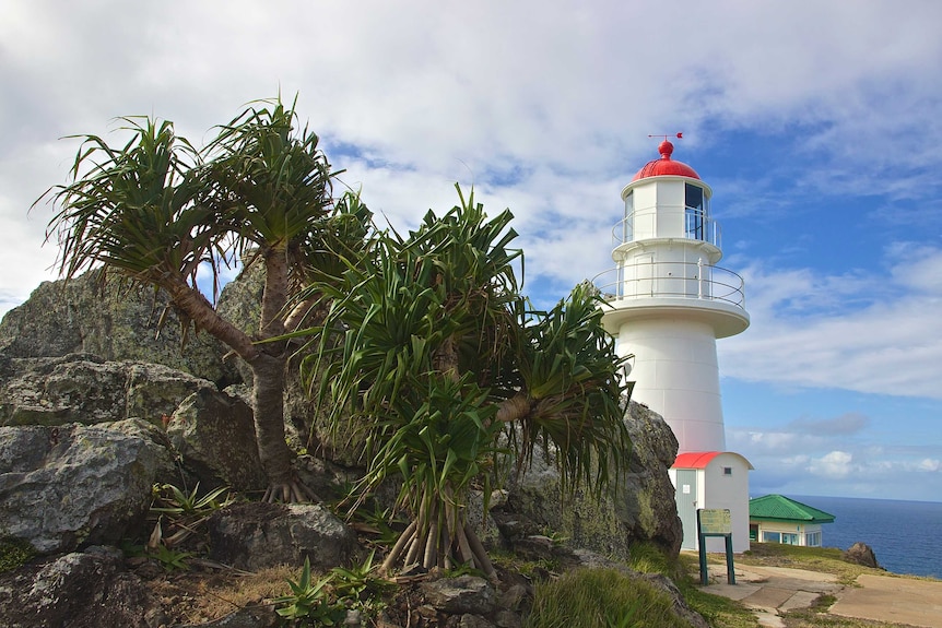 A white lighthouse with a red top stands nestled into boulders with vegetation growing on them.