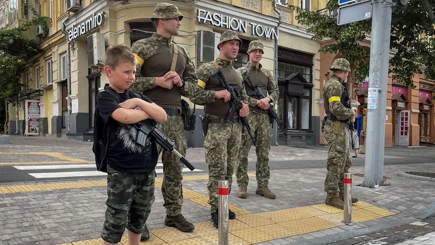 A boy with a toy machine gun stands near Ukrainian servicemen on a street.