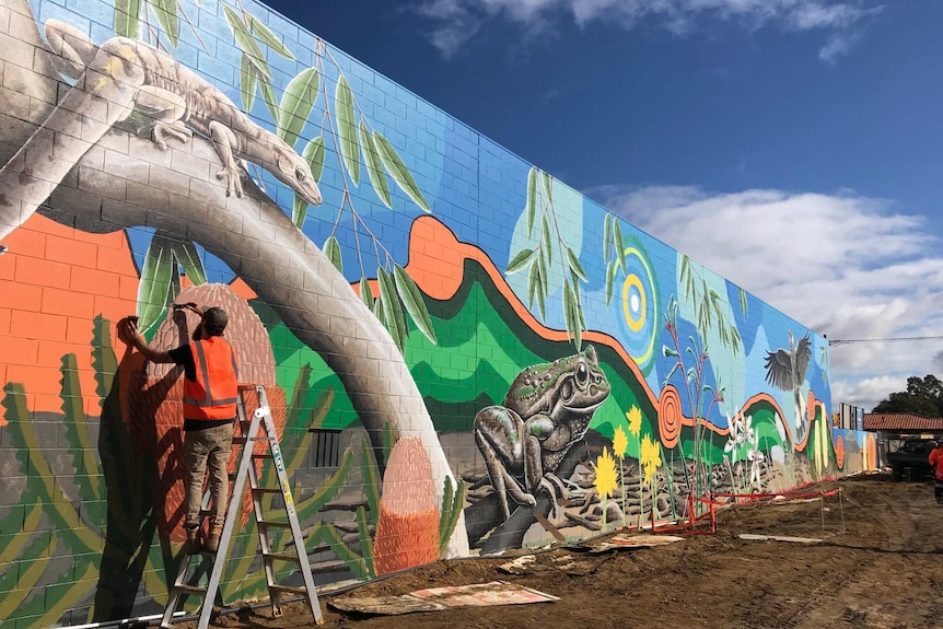 A man in hi-vis on a ladder paints a mural on a brick wall