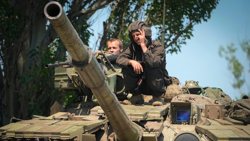 A soldier flashes the victory sign atop a tank while another looks ahead.