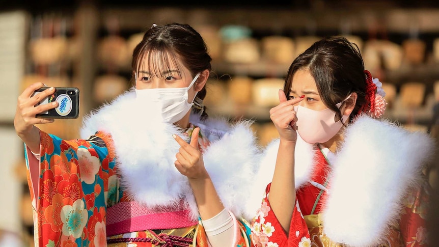 Two women wearing masks and kimons hold a phone to take a selfie.