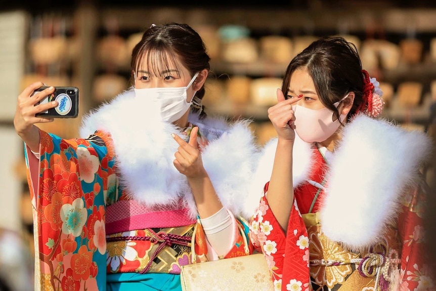 Two women wearing masks and kimons hold a phone to take a selfie.