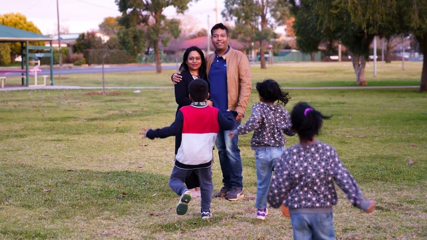 A man his wife smile at the camera while three children run towards them
