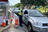 A police officer at a check point speaks to someone in a car, with other cars line up behind