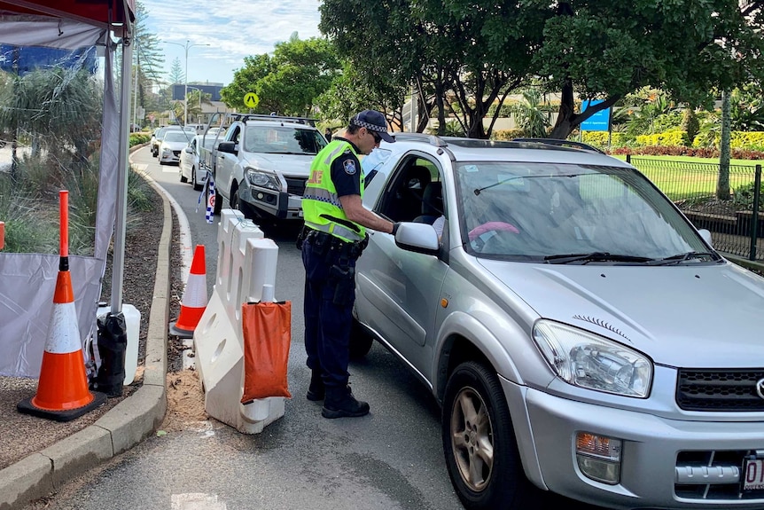 A police officer at a check point speaks to someone in a car, with other cars line up behind