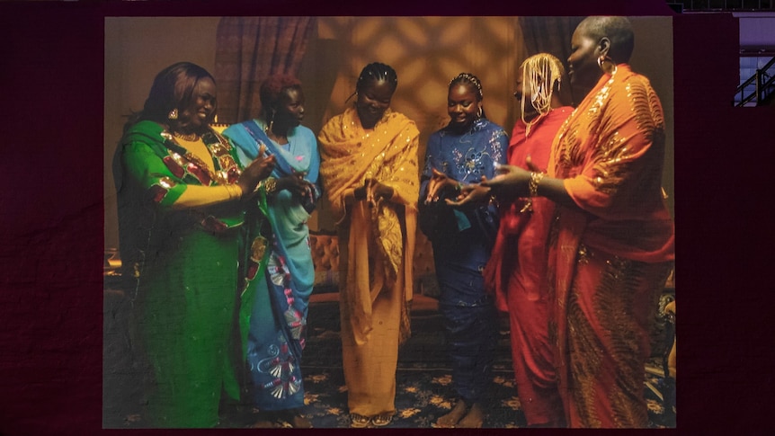 A video projection of six African women standing in bright dresses and smiling, their hands pressed together as if mid-clap