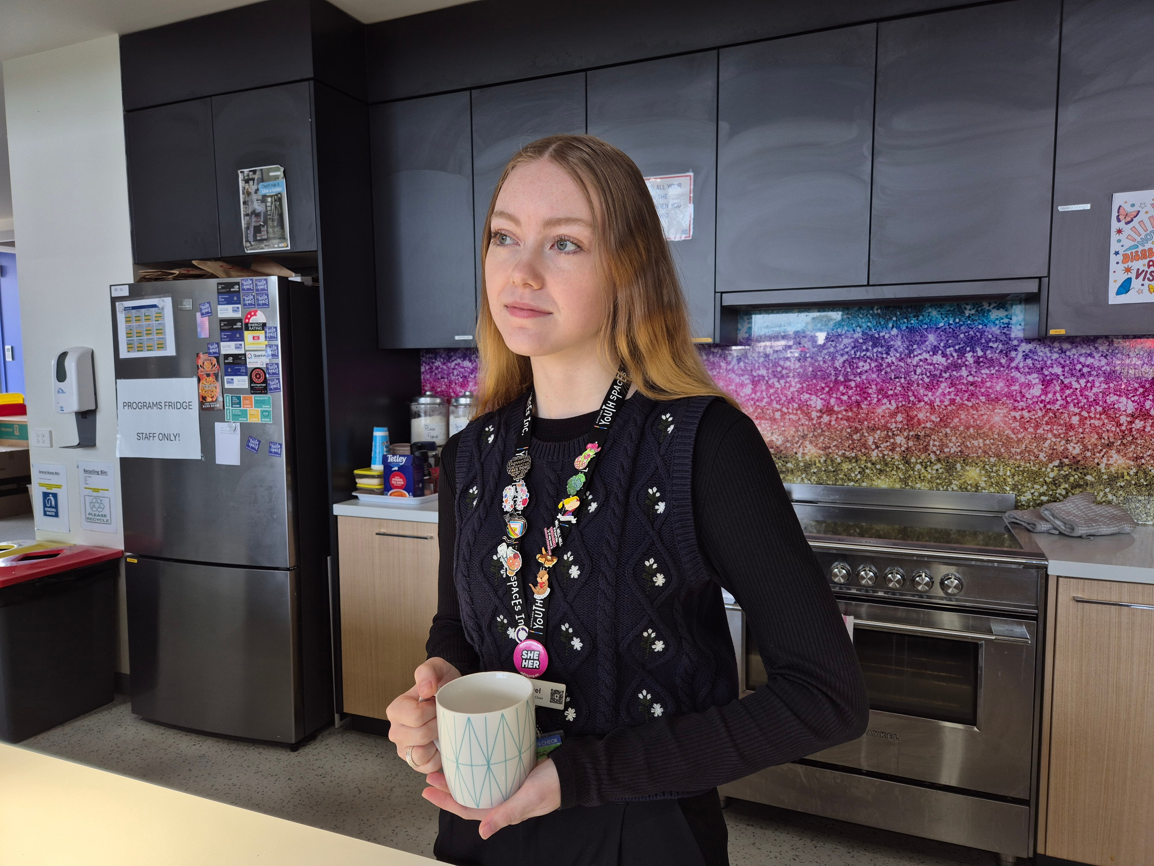A young woman with strawberry blonde hair in a dark top holds a mug in a kitchen, looking thoughtful