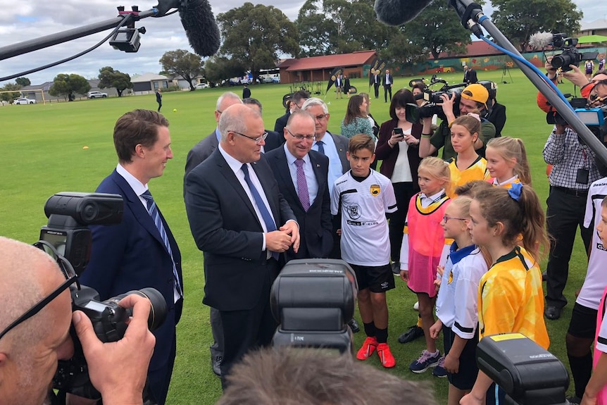 Politicians in suits stand in front of cameras and children in a football field