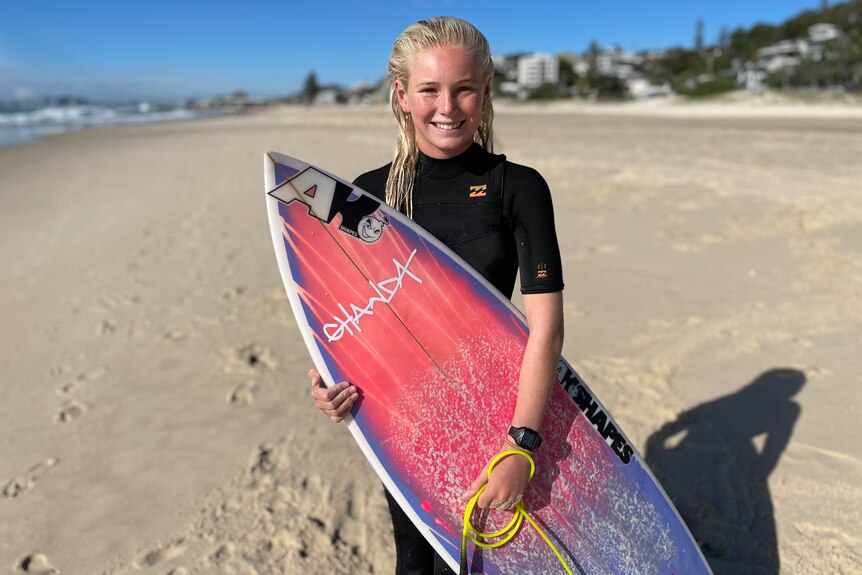 a young female surfer in a wetsuit standing on the beach with her surfboard