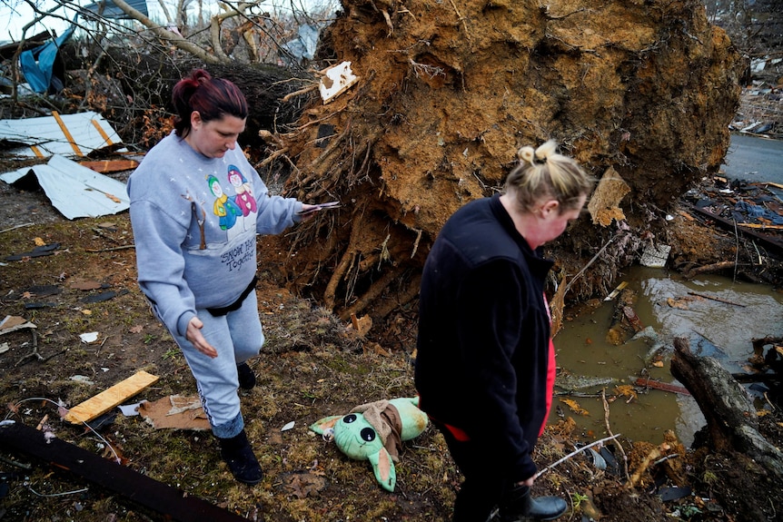 Two women search of belongings, uprooted tree in background.