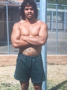 A man, wearing shorts, footy socks and thongs stands in jail