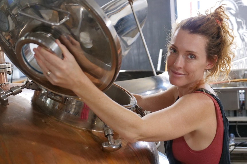 A woman with orange hair tied up in a bun holding the lid open on a copper still while smiling.