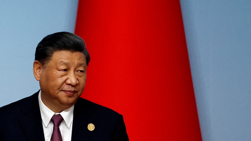 A close up of Xi Jinping wearing a suit looking over his shoulder in front of a flag.
