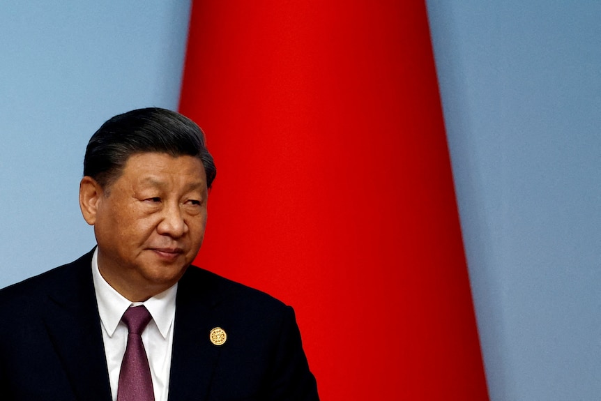 A close up of Xi Jinping wearing a suit looking over his shoulder in front of a flag.