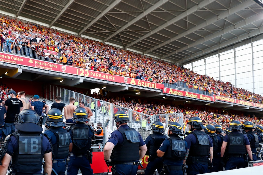 Une ligne de policiers anti-émeute surveille un stand complet de supporters vêtus de rouge et de jaune