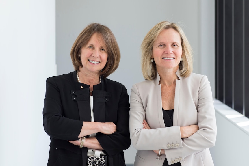 Suzanne Crowe and Sharon Lewin smile as they stand beside one another in a corridor.