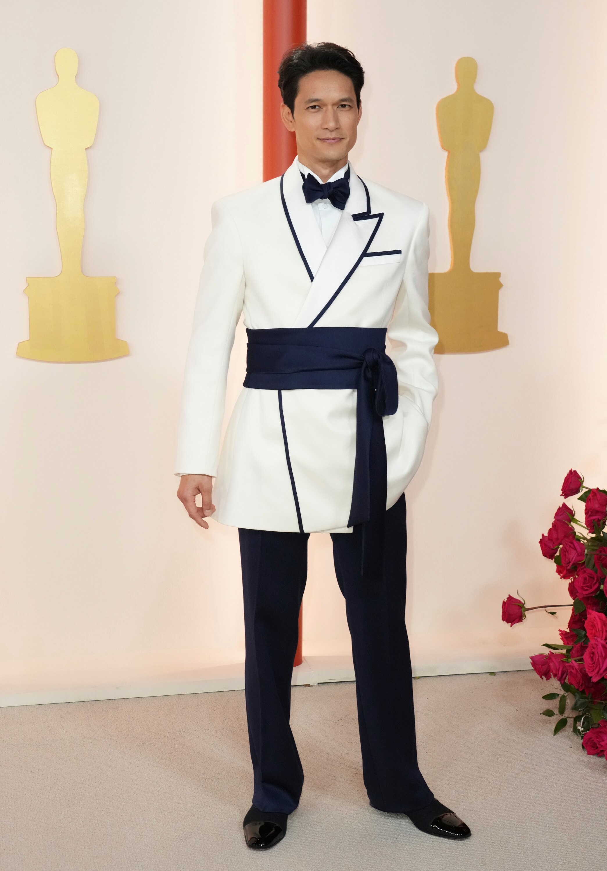 Harry Shum Jr wearing a white long suit jacket with a black bow tie, a black sash around his middle and black trousers. 