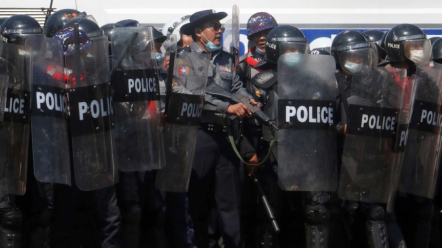 A police officer holds a weapon while standing among a row of riot police who hold up clear shields.