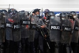 A police officer holds a weapon while standing among a row of riot police who hold up clear shields.