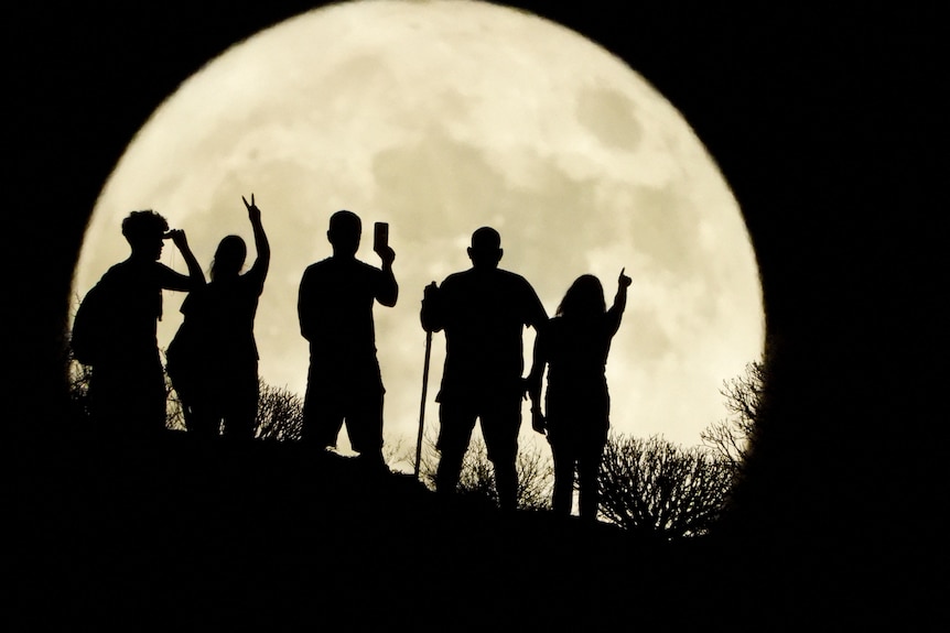Five people stand in front of a large, yellow full moon on top of a hill